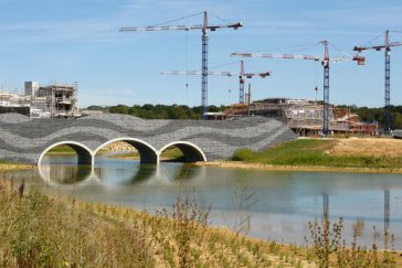 Au premier plan : une rivière est surplombée par un pont. Au second plan : plusieurs grues du chantier de Villages Nature Paris