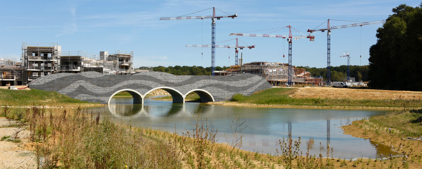 Au premier plan : une rivière est surplombée par un pont. Au second plan : plusieurs grues du chantier de Villages Nature Paris