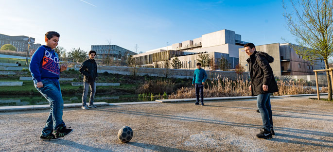 Crédit : ©Epamarne / Architectes : PC Bontempi - Hertenberger et Vitry / Paysagiste : Atelier de l'Ile / Photo : Antoine Meyssonier Photographie de quatre adolescents jouant au football aux abords du collège et lycée international de l'est parisien à Noisy-Bry