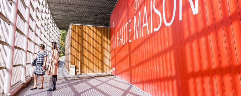 Deux passants regardent au loin, derrière le grillage, dans leurs dos, on aperçoit la façade de la Haute Maison, bâtiment présent à la Cité Descartes