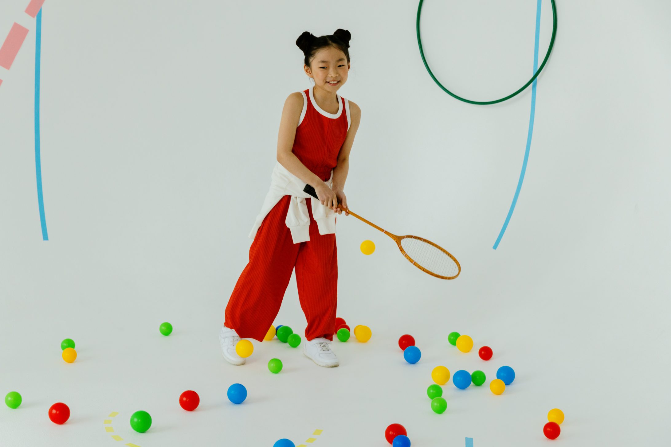 Une fille sur fond blanc, avec une raquette de badminton à la main et des boules au sol. 